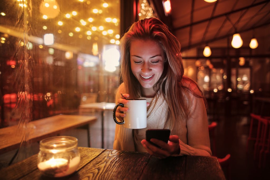 Woman smiling while holding coffee and using phone in warm caf&eacute; ambiance.